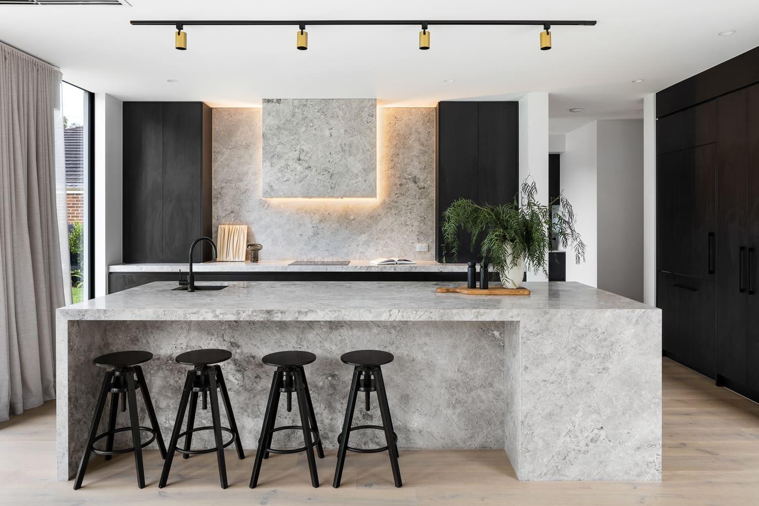 A contemporary kitchen with natural stone island bench and splash back and stylish black stools.